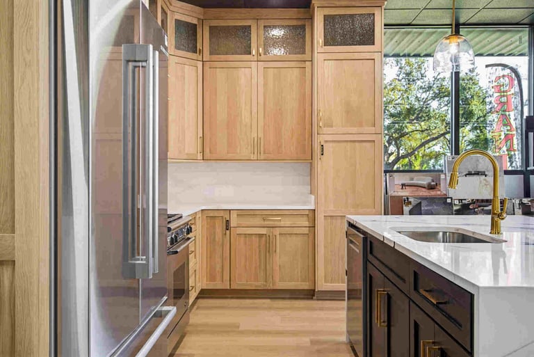 Modern kitchen with light wood cabinetry, stainless steel refrigerator, white quartz countertops, black island, brass faucet, and large windows overlooking exterior scenery.
