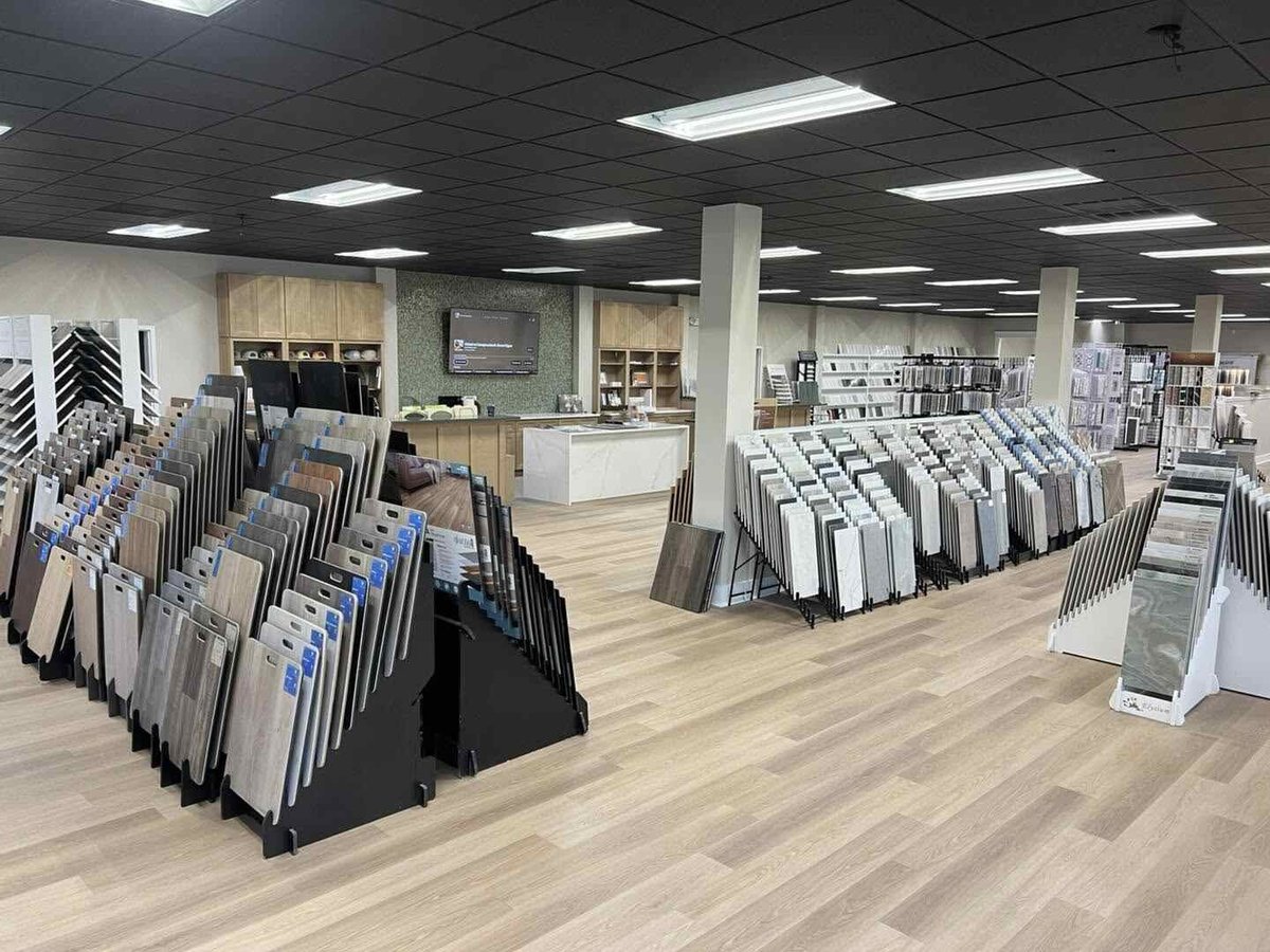 Modern showroom with flooring and tile samples displayed on racks, sales counter, and product shelving in bright, open space.
