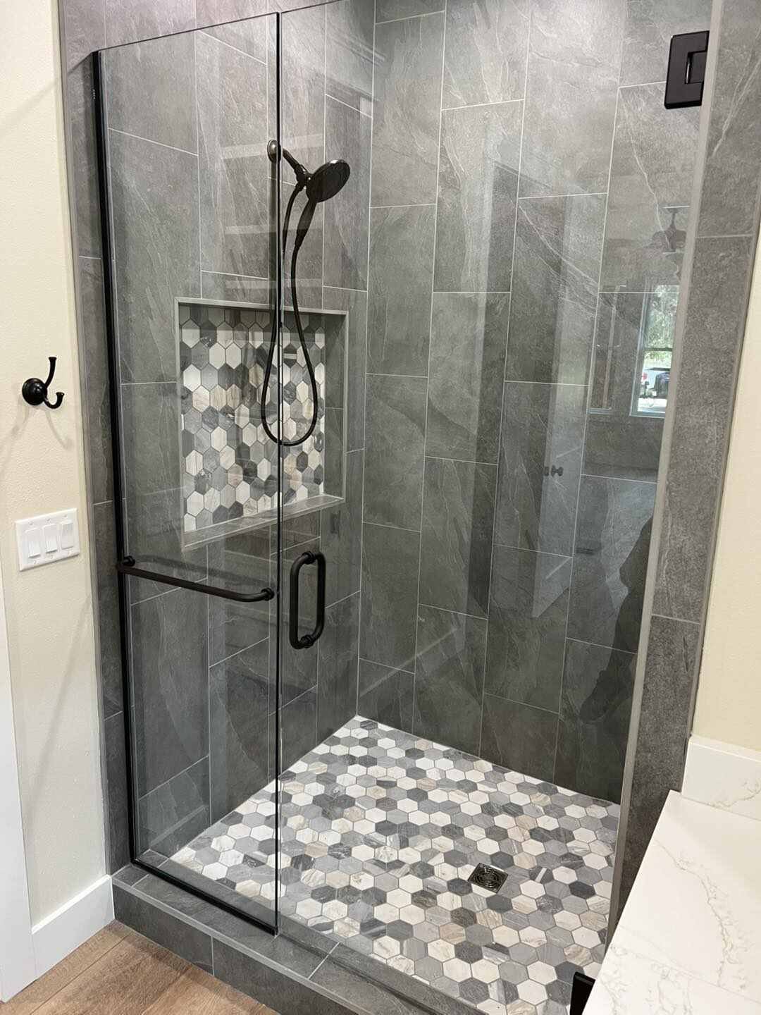 Modern gray tile shower enclosure with glass doors, black fixtures, and hexagonal mosaic floor
