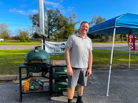 Man standing next to a green egg-shaped outdoor grill in a park with trees and a blue canopy tent in the background