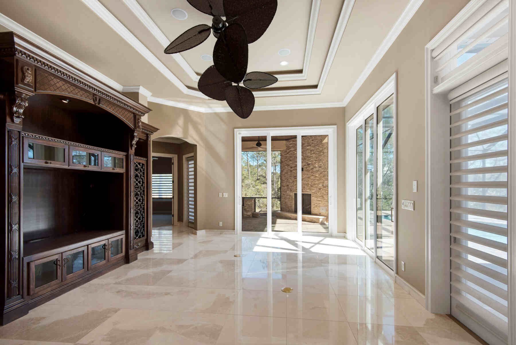 Modern open-concept living room with polished marble floors, dark wood entertainment center, ceiling fan, and glass doors opening to outdoor patio