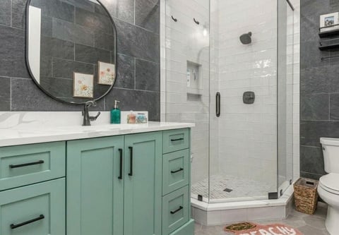 Modern bathroom with turquoise vanity, black round mirror, gray tile walls, and glass-enclosed walk-in shower with black fixtures