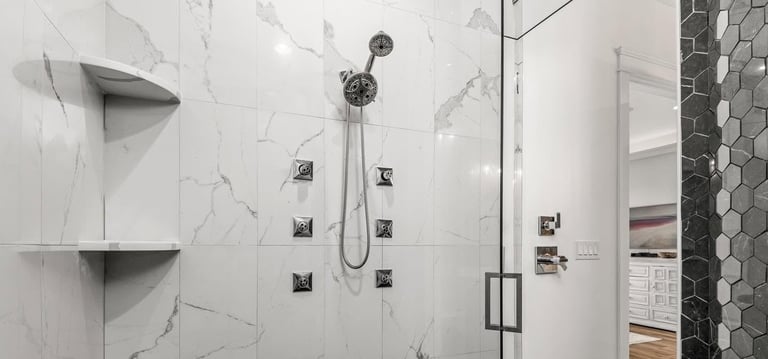 Modern bathroom shower wall with marble-look tiles, rainfall showerhead, body jets, and stainless steel fixtures with glass enclosure