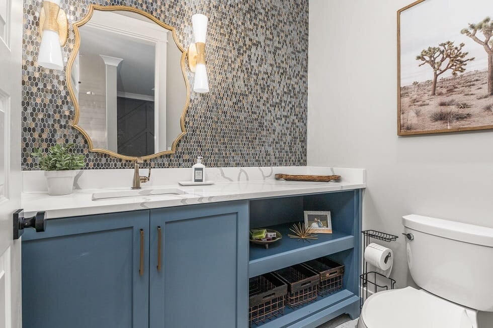 Modern bathroom with blue vanity, white marble countertop, round mirror with gold frame, patterned tile wall, and toilet