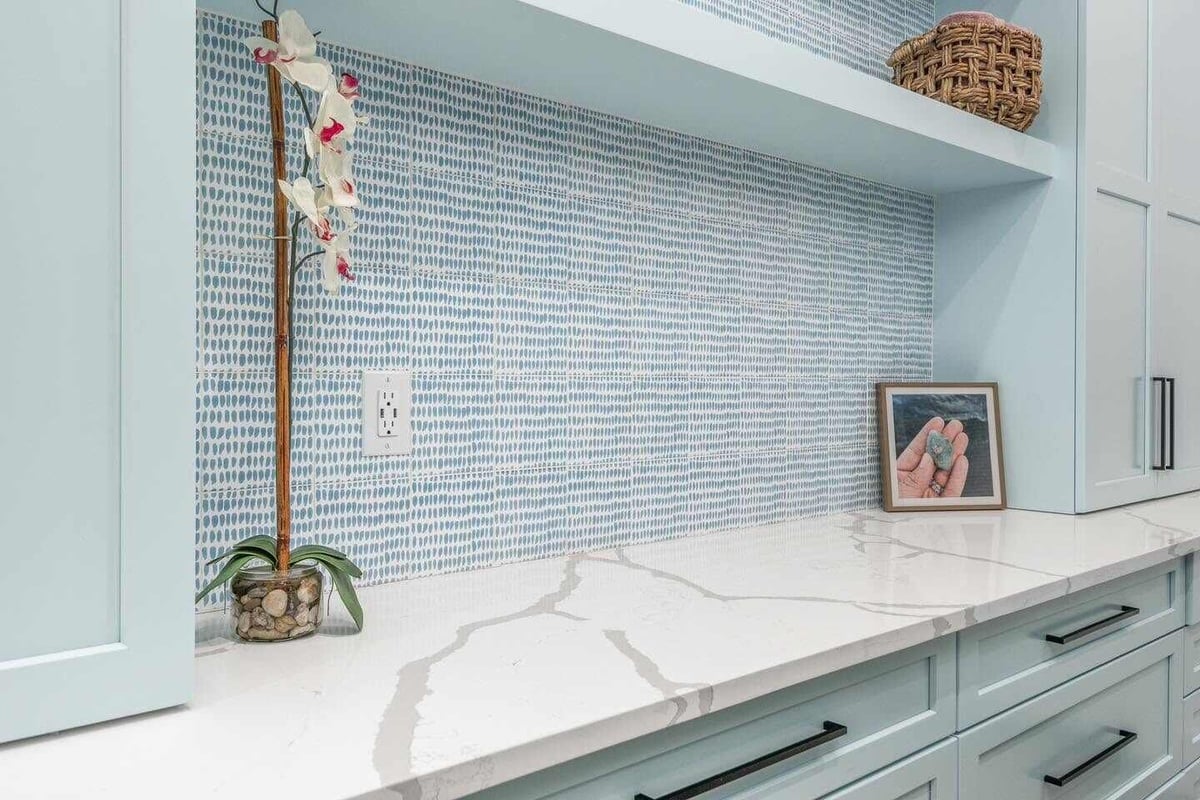 Modern kitchen countertop with light green cabinetry, white marble backsplash, and blue tile accent wall with decorative items and art.
