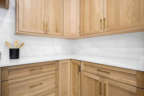 Modern kitchen corner with natural wood cabinetry, white marble countertops and backsplash, brass hardware, and a black utensil holder