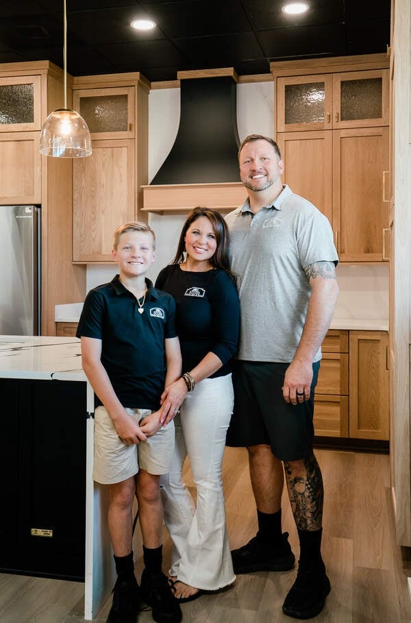 Three people posing in a modern kitchen with wood cabinetry and stainless steel appliances