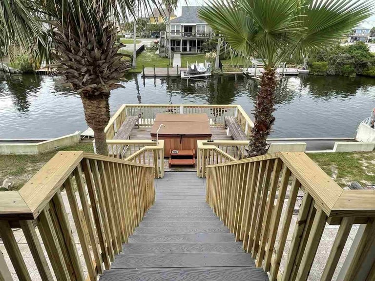 Wooden boardwalk with railings leading to a waterfront deck, flanked by palm trees with waterfront homes and boats visible across the canal