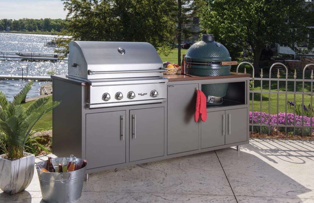 Outdoor kitchen setup with stainless steel grill, green egg smoker, and storage cabinets on a patio near water and landscaping