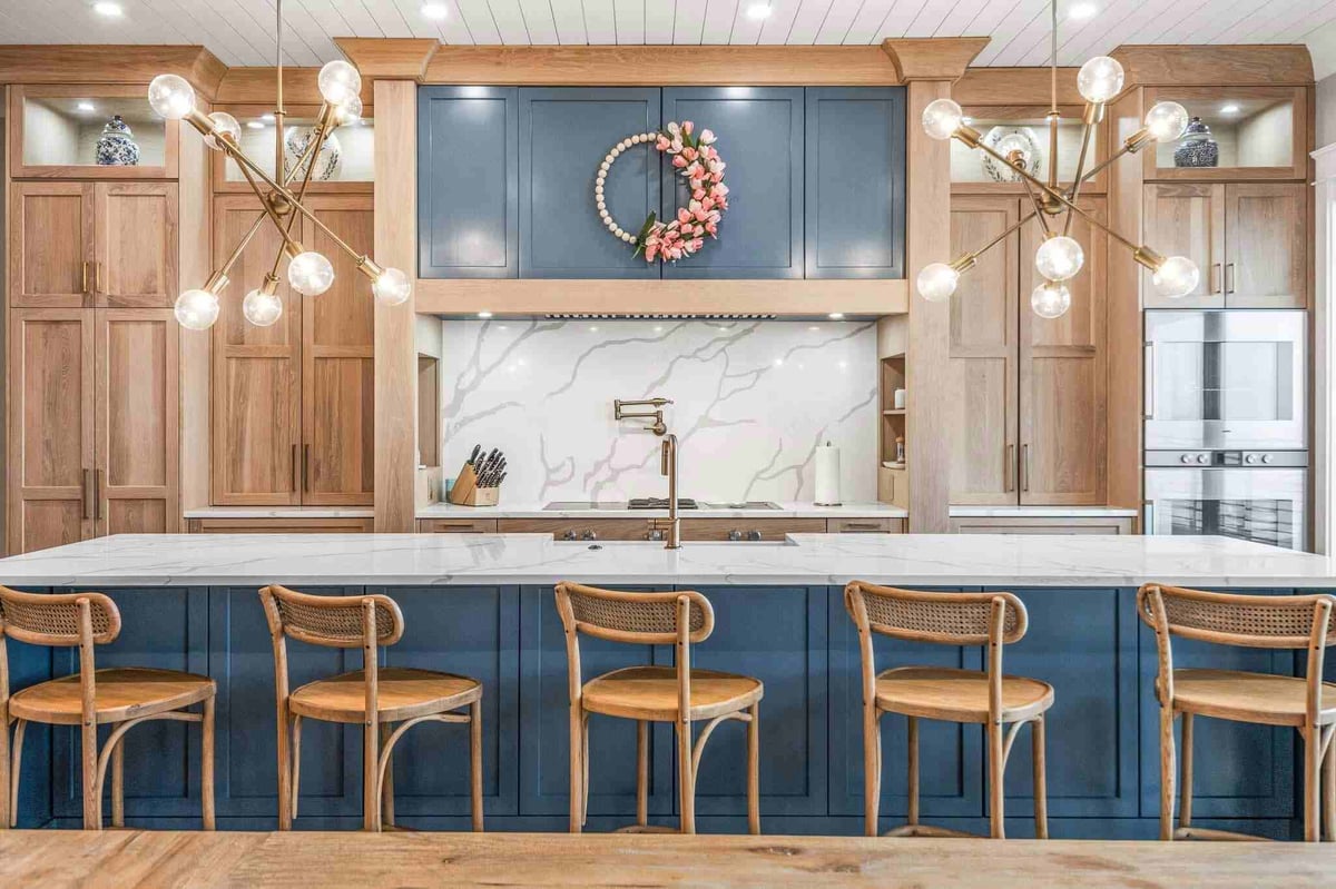 Modern kitchen with blue cabinetry, marble backsplash, wooden bar stools, pendant lights, and a floral wreath above the stove hood