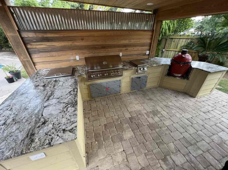 Covered outdoor kitchen with marble countertops, built-in stainless steel appliances, and red grill on brick patio
