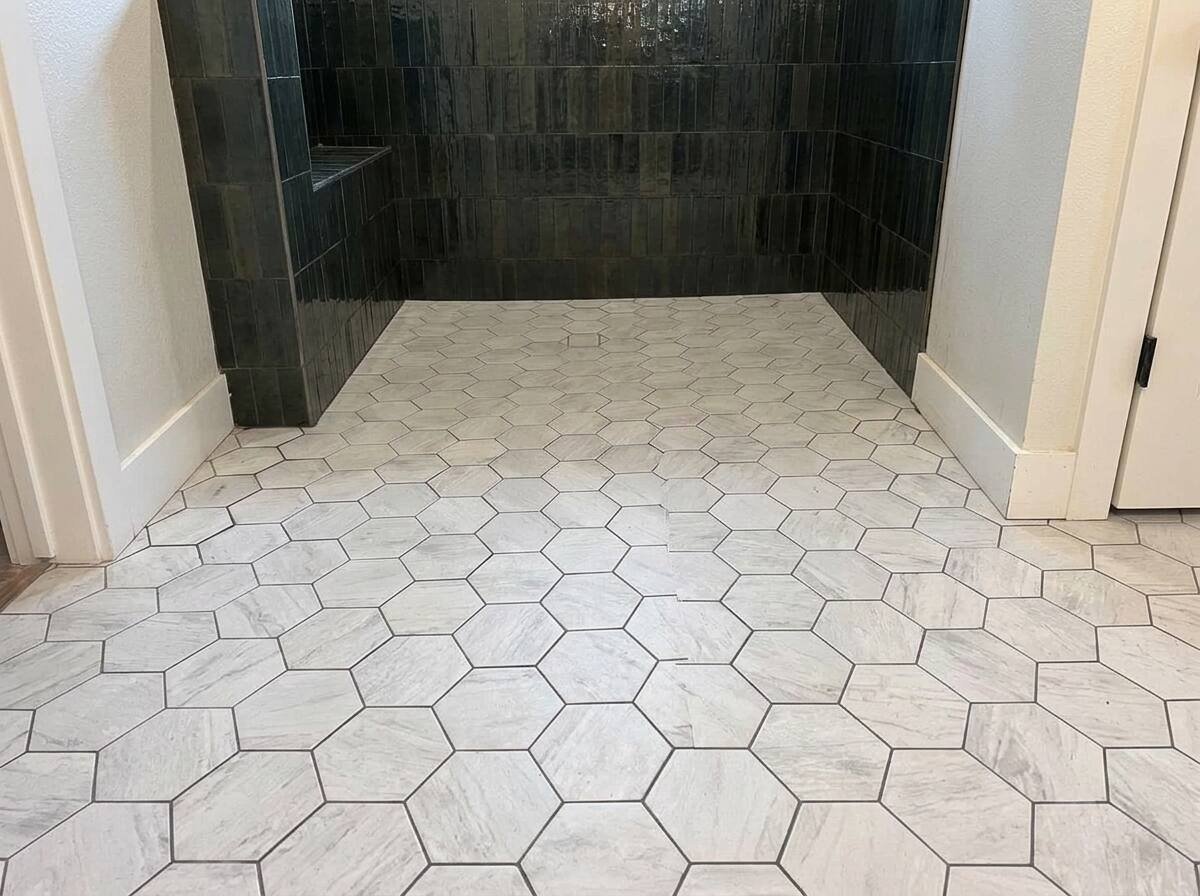 Modern bathroom with hexagonal tile flooring in light marble tones, black tiled shower enclosure at back, and white doorframes on sides